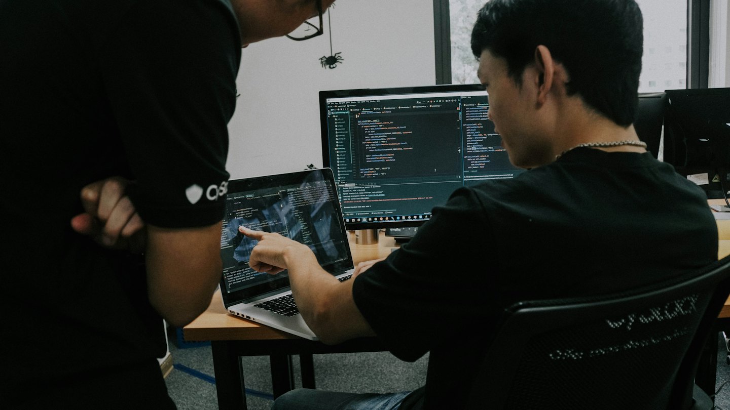 Two men review code on a laptop in an office.